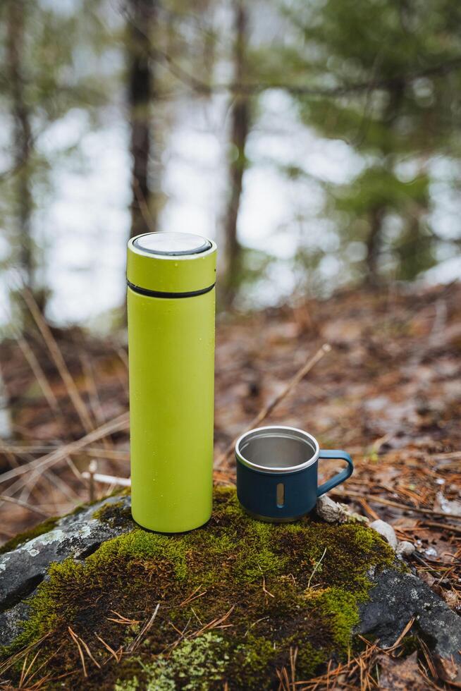 A close-up of a thermos stands on a stone next to a mug made of metal, camping utensils. photo