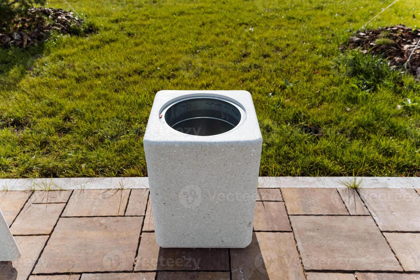 In front of a grassy field, there is a white trash can placed on a brick patio, creating a contrast between the patios composition and the natural greenery surrounding it photo