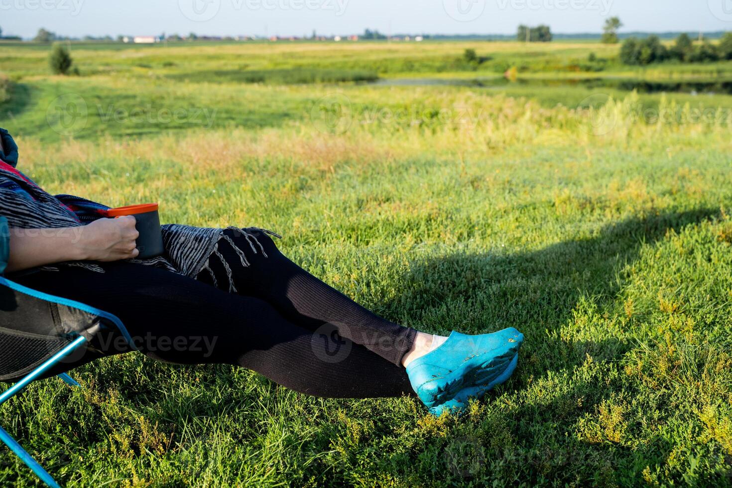 In a serene landscape, a woman is comfortably sitting in a chair with her feet propped up, surrounded by a vibrant field filled with lush grass and under a clear blue sky photo