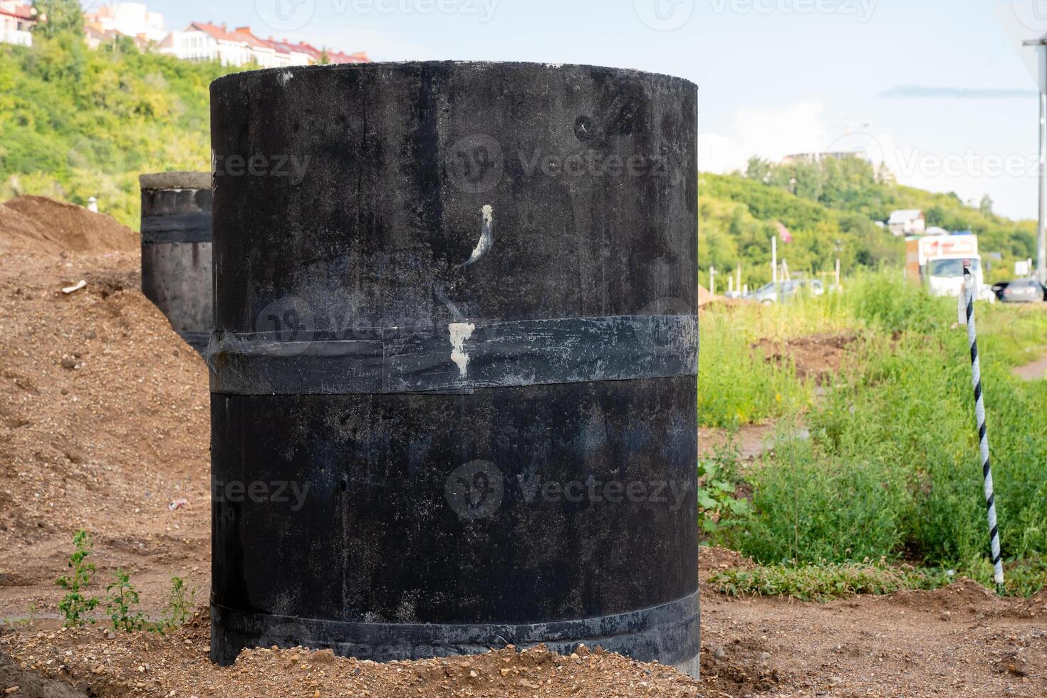 In the center of a wide dirt field, there is a large black cylinder prominently sitting, making it the focal point of the surroundings with grass and trees in the background photo