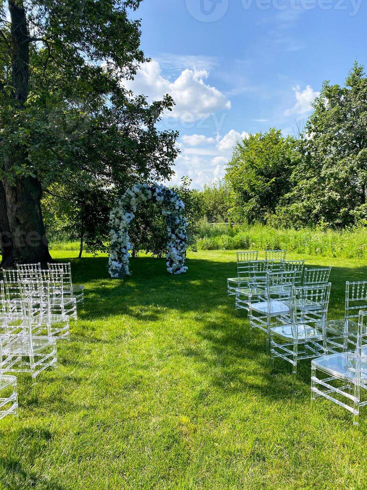 A row of white chairs is arranged in a grassy field for a wedding ceremony beneath the expansive sky. The setting includes elements like water, clouds, plants, and grass photo