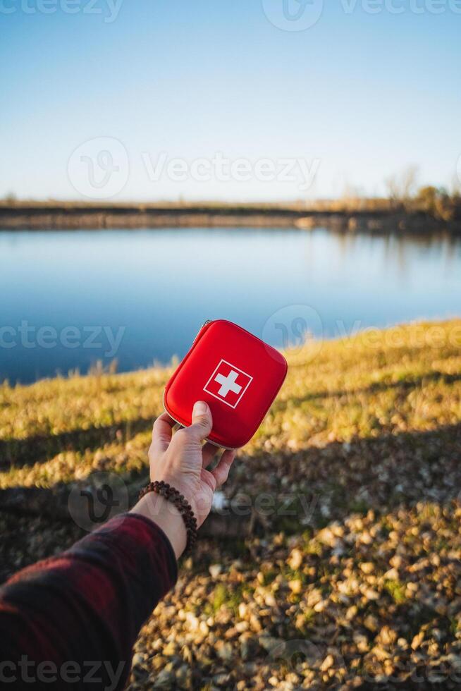 A cheerful person beams with joy, holding a bright red first aid kit by a tranquil lake, surrounded by lush green grass, under a stunning clear blue sky in warm sunlight photo