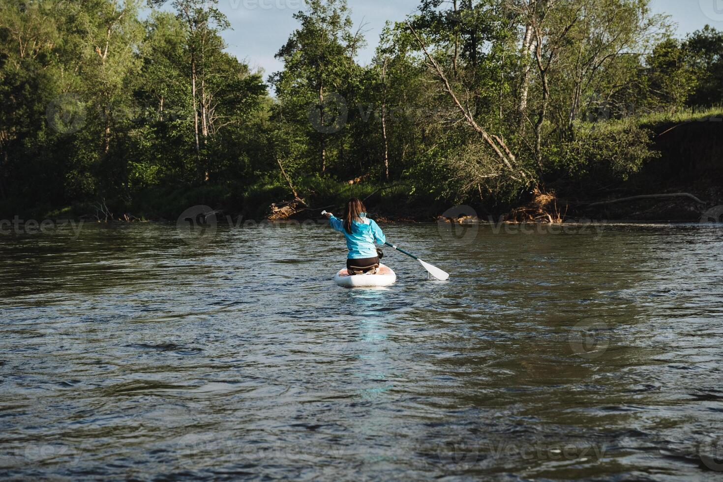 A woman peacefully paddles on a tranquil river, admiring natural beauty under a clear sky. The serene setting creates a calming atmosphere, surrounded by soothing nature photo