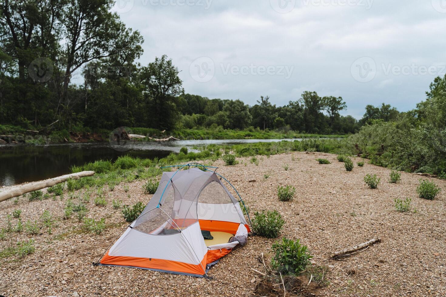 A tent is pitched on a sandy beach by a river, surrounded by natural landscape. The serene setting offers a peaceful escape with water, trees, grass, and a clear sky above photo