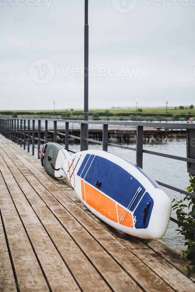 On a cloudy day, a paddleboard rests on a wooden dock by the waterfront, creating a serene coastal scene with marine elements. The setting exudes tranquility and beauty photo
