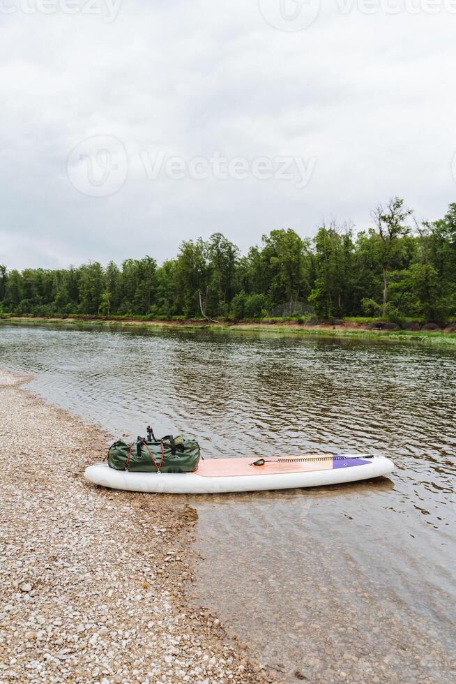 A serene scene with a paddle board resting by a calm riverbank under cloudy skies, perfectly harmonizing with its surroundings to convey a sense of peace and connection with nature photo