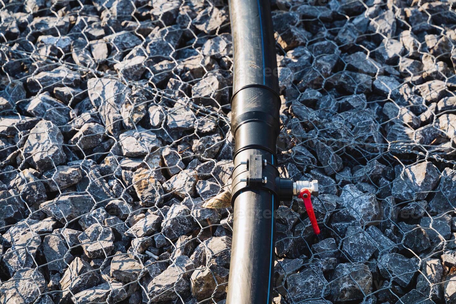 A sturdy black pipe featuring a bright red valve is positioned on top of an uneven pile of rocks, creating an interesting contrast between the industrial and natural elements around it photo