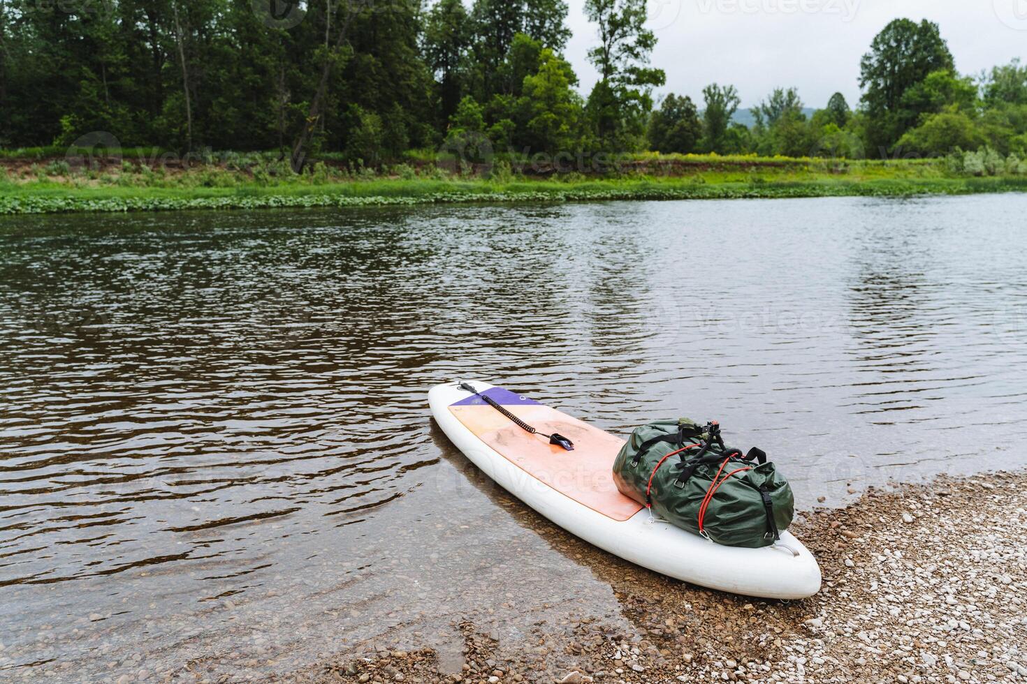 Indulge in the tranquility of paddleboarding on a tranquil riverbank, surrounded by lush greenery and calm waters under an overcast sky, with camping gear for an outdoor adventure photo