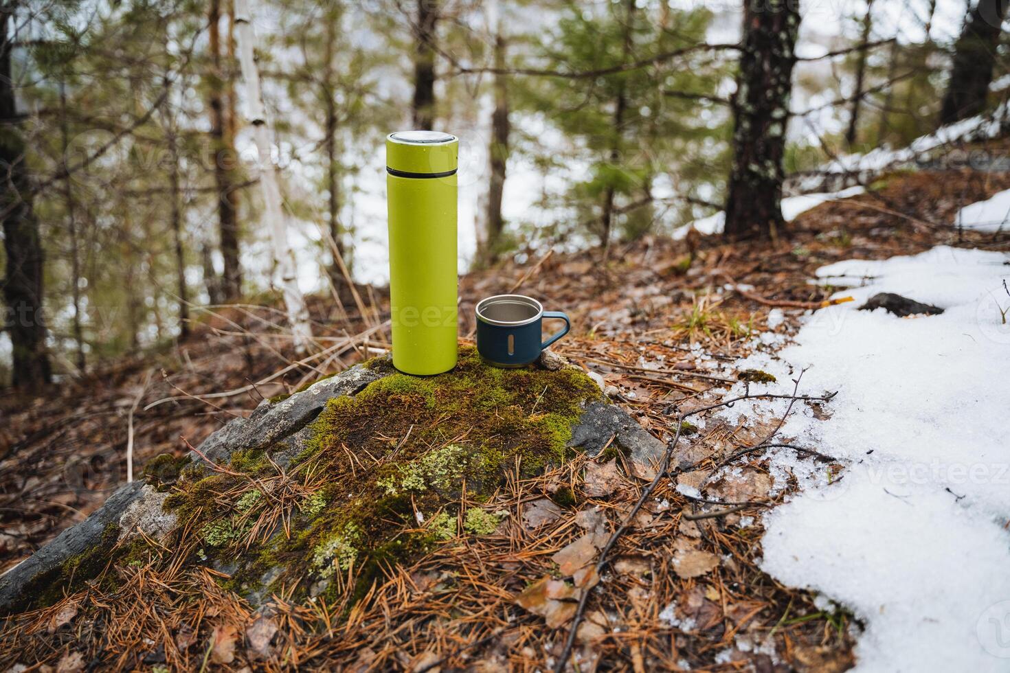 A light green thermos stands on a stone in the forest next to a metal mug, a concept of tourist utensils for trekking and walking. photo