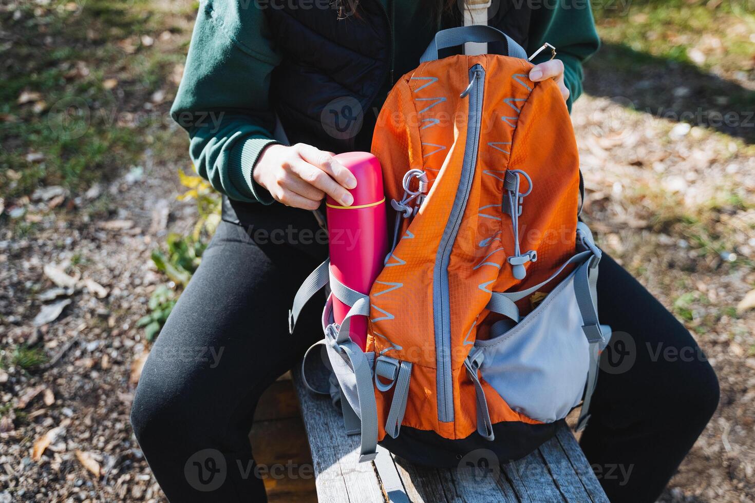 A woman sits on a park bench with a bright orange backpack next to her and a pink water bottle in hand, surrounded by a stunning outdoor setting of greenery and vibrant colors photo