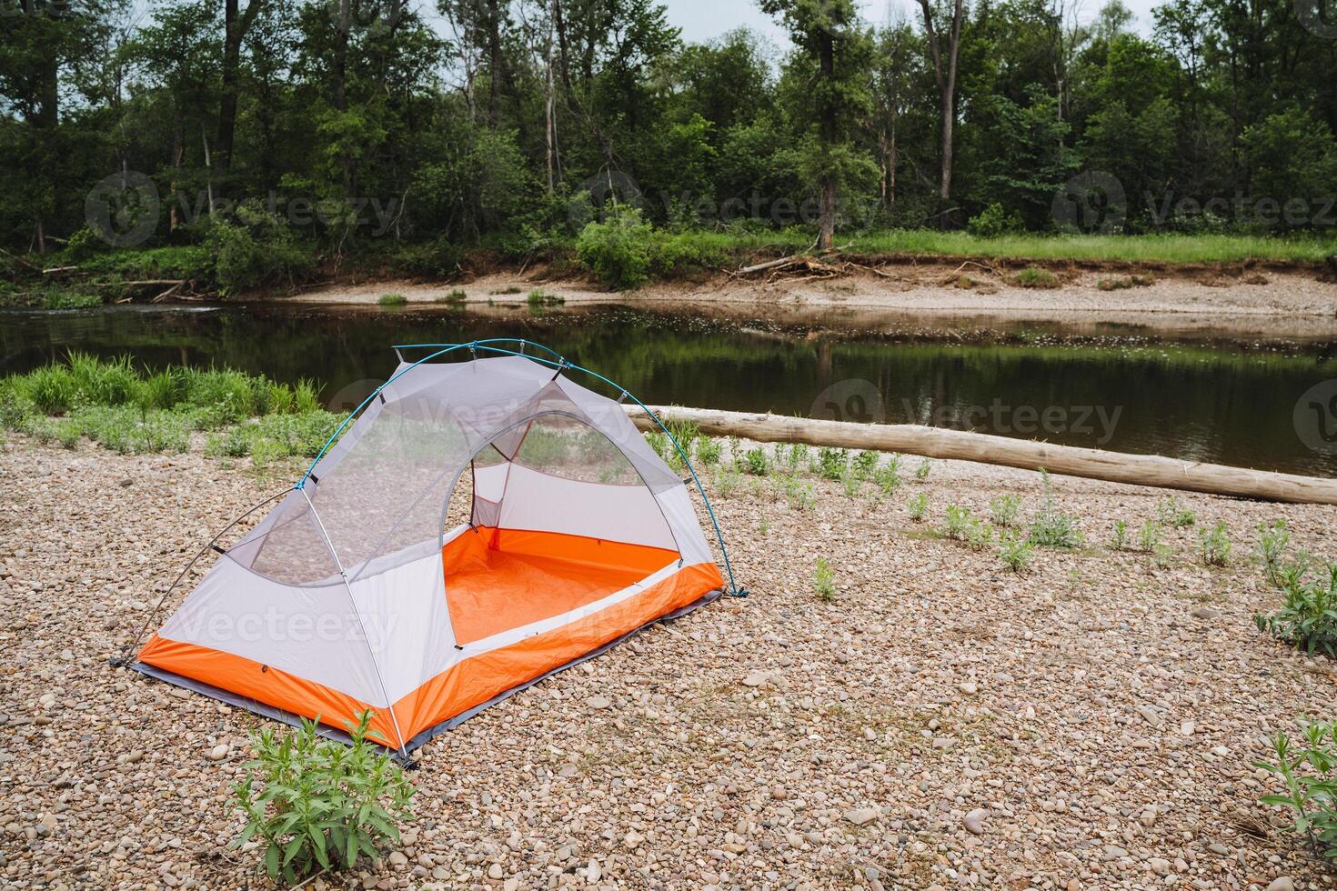 Experience the tranquility of camping by a peaceful river, with a tent pitched on the rocky bank in a lush forest ideal for relaxing and connecting with nature photo