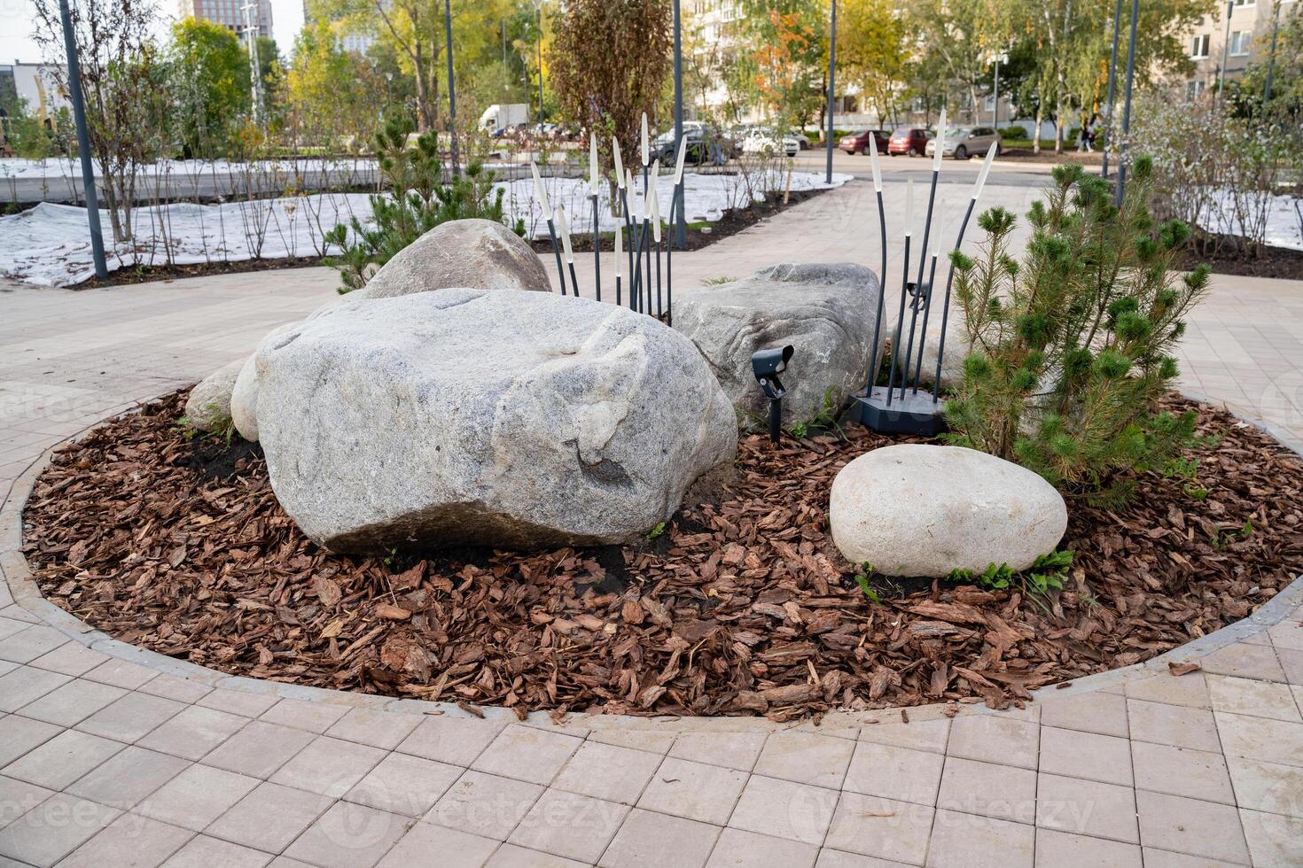 In the park, a large rock is prominently sitting in a welldefined circle of mulch. This area contrasts with the surrounding grass and various other plants within the landscape photo