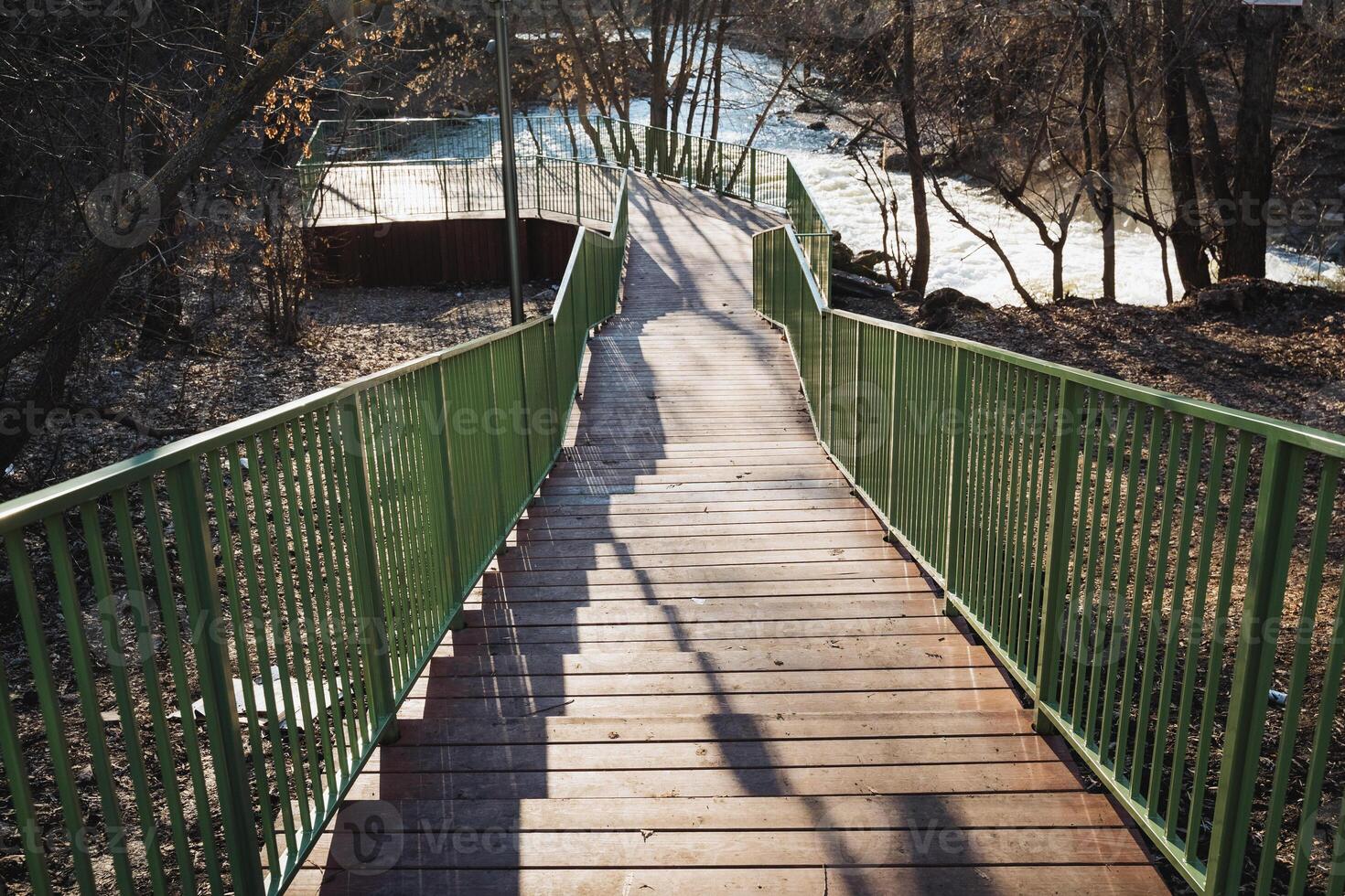 A beautifully scenic wooden footbridge gracefully stretches over a serene, calm river, surrounded by vibrant nature in the soft morning light, offering a perfect escape for all photo