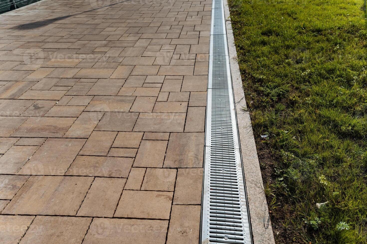 There exists a drain situated on the sidewalk that runs parallel to a grassy patch, which is adorned with vibrant lush green grass and various landscape features enhancing its beauty photo