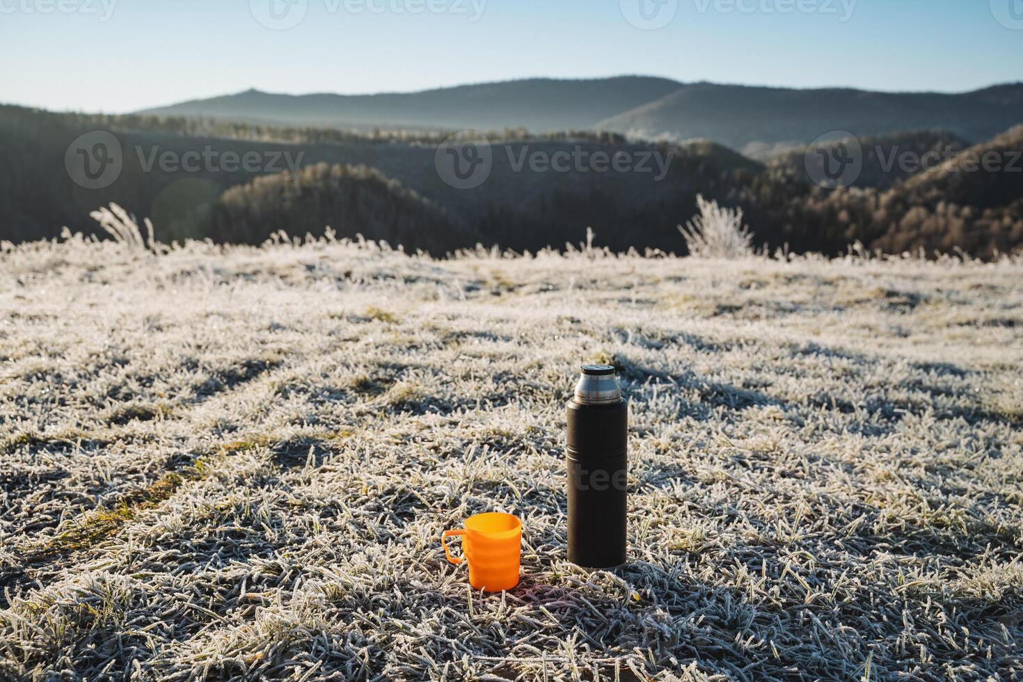 Black thermos stands on the grass against the background of mountains, hot drink in thermal bottle, orange mug, drinking hot tea outdoors in winter. photo
