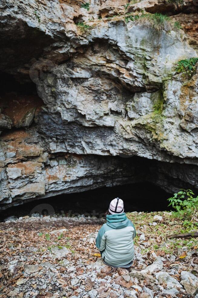A person is comfortably sitting on the ground in front of a large, mysterious cave, surrounded by various natural elements and an intriguing landscape that captures the imagination photo