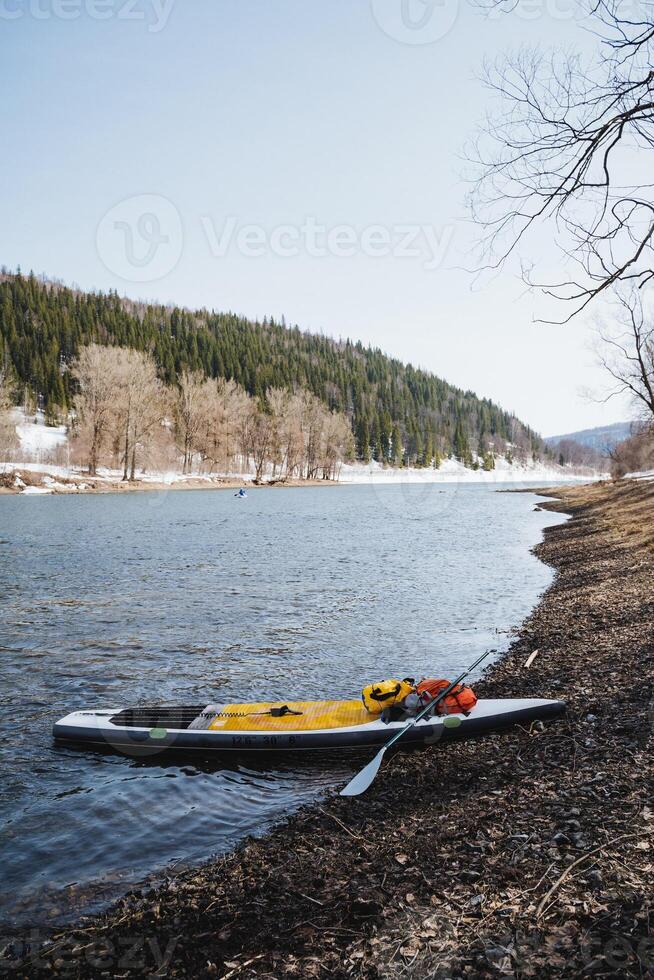 A SUP board stands by the bank on the river against the background of mountains, a tourist trip on the water on a board with a paddle, spring rafting. photo