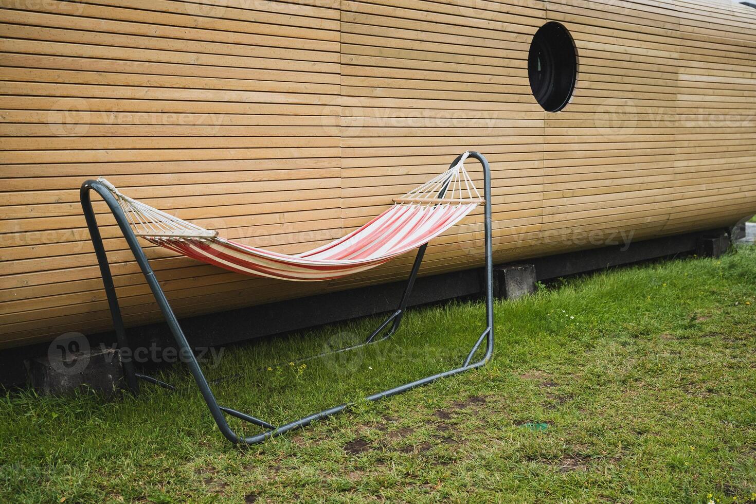 In a serene outdoor setting, a lively hammock on a metal frame stands next to wooden siding, offering a tranquil escape in a lush grassy yard for relaxation and leisure photo
