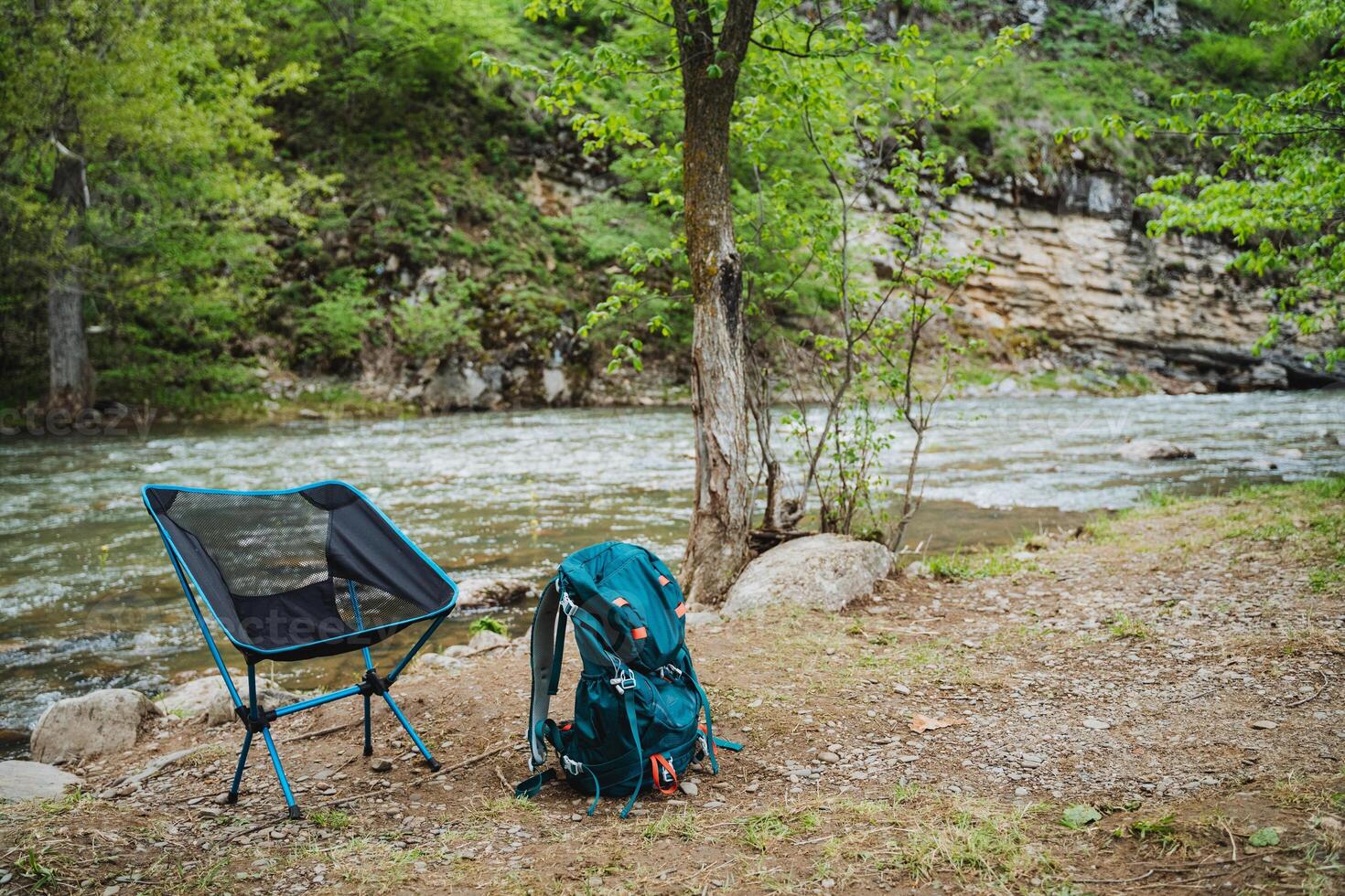 Experience a serene camping scene by a lush riverside in a forest with a backpack and chair. Explore nature, relax by the water, and enjoy the scenic landscape photo