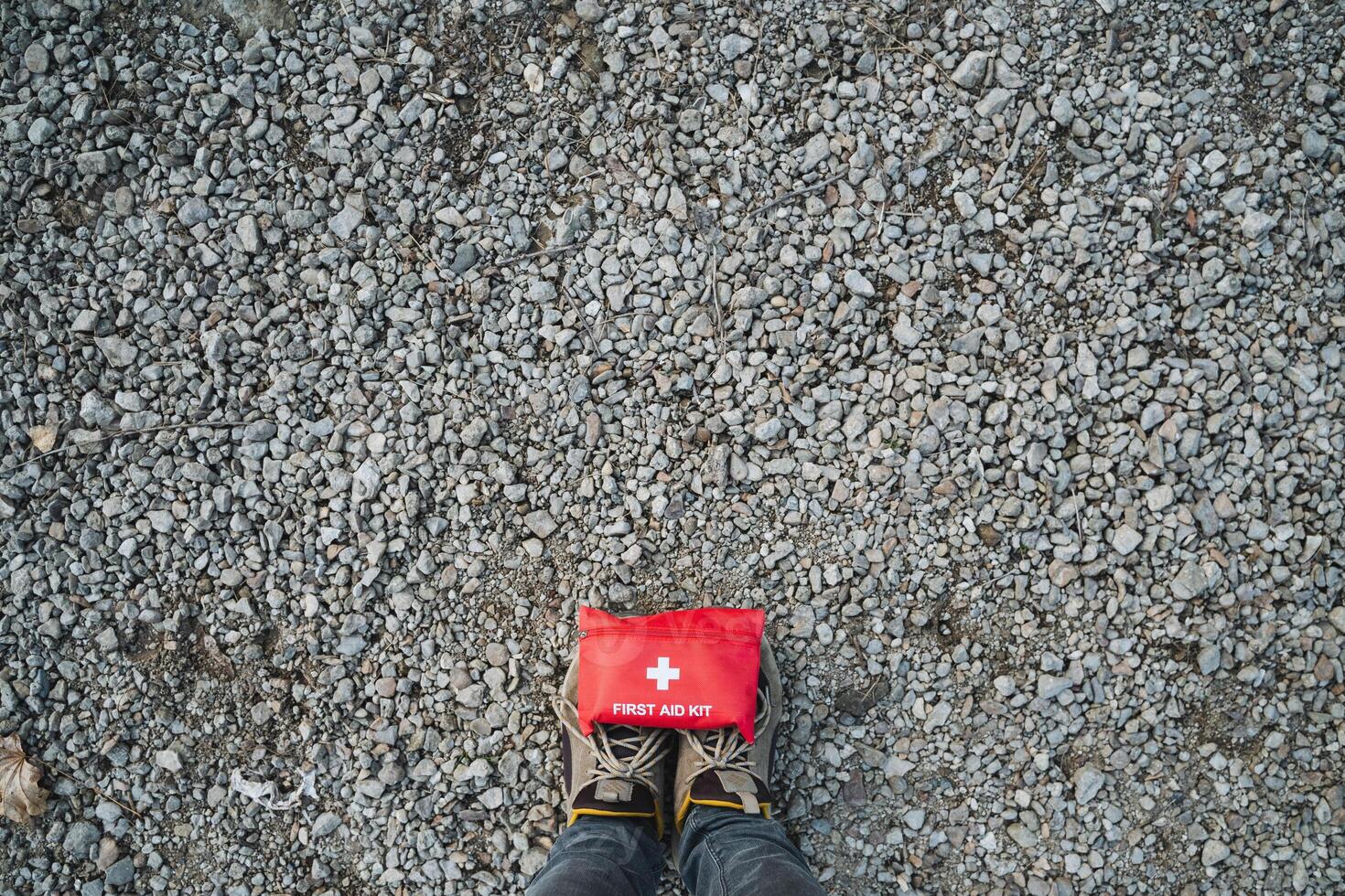 An Emergency First Aid Kit is placed on a gravel surface, paired with sturdy hiking boots, ideal for outdoor exploration and prevention photo