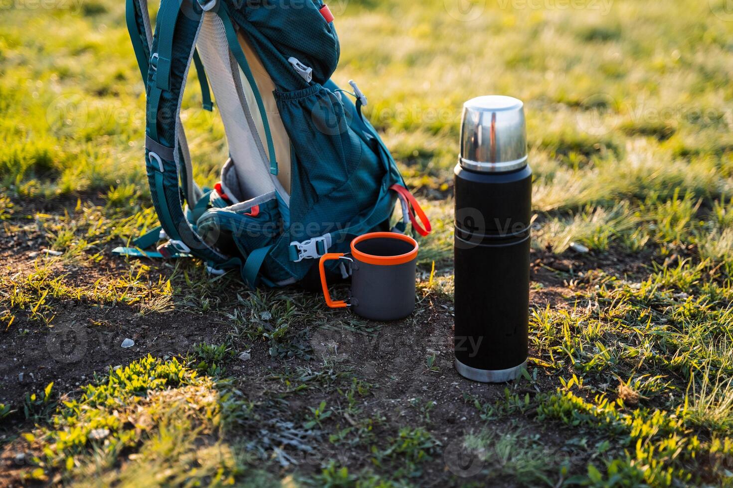 A water bottle lies on the ground next to a thermos and a mug in the natural environment. Nearby, an automotive tire, plastic bottle, and camera lens are scattered among the grass and trees photo
