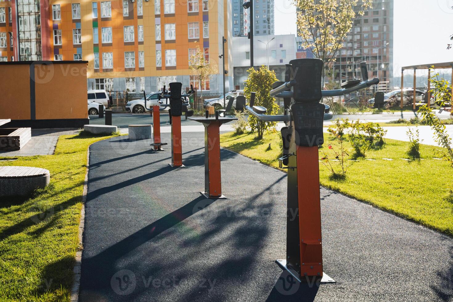 In a public park area, a row of diverse exercise equipment is arranged, and a nearby building can be seen in the background, which adds to the surrounding urban landscape photo