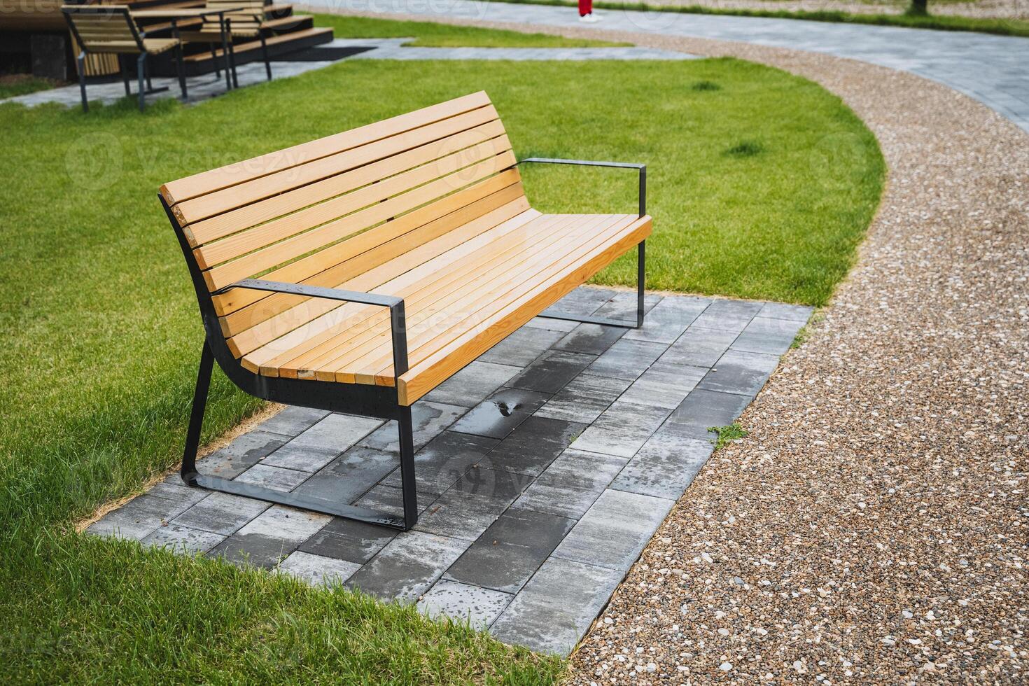 In a peaceful park, a basic wooden bench sits on a red brick path amidst green grass and a stunning natural backdrop, providing a serene spot to relax and admire the surroundings photo