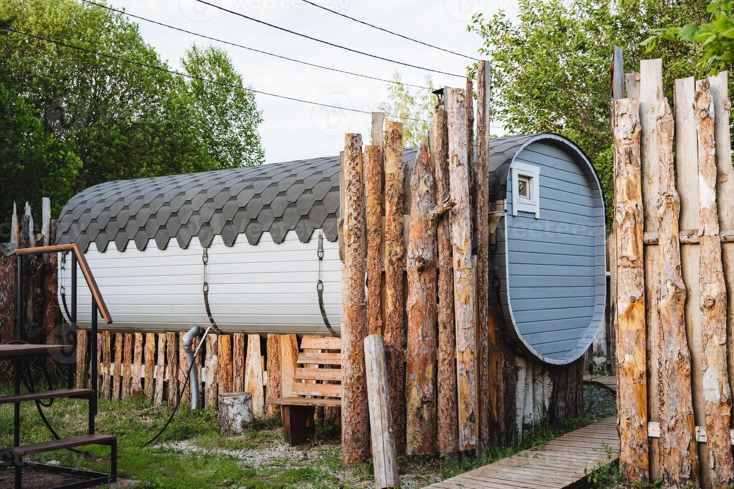 A barrelshaped structure is nestled within the scenery, enclosed by a wooden fence, effortlessly fitting in with the lush landscape featuring trees, grass, and a house topped with a roof photo