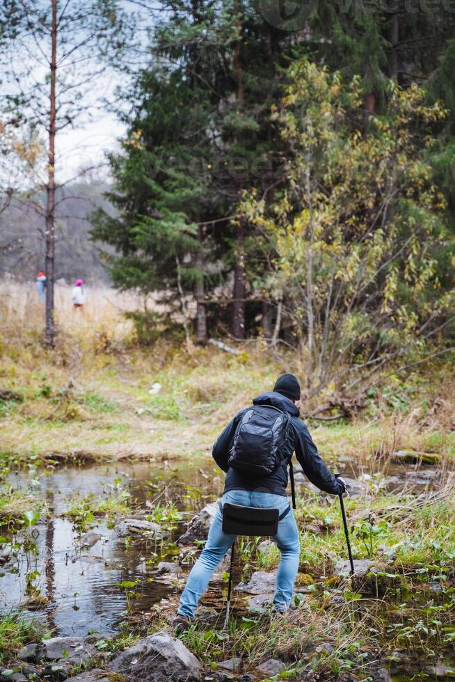 A man, who is carrying a backpack, is carefully crossing a stream that runs through the beautiful woods filled with trees and grass, surrounded by a peaceful natural landscape photo