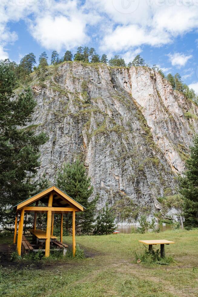 A serene and beautiful wooden gazebo stands in lush green grass, set against a majestic mountain backdrop. The scenery includes clouds, sky, trees, and a natural highland landscape photo