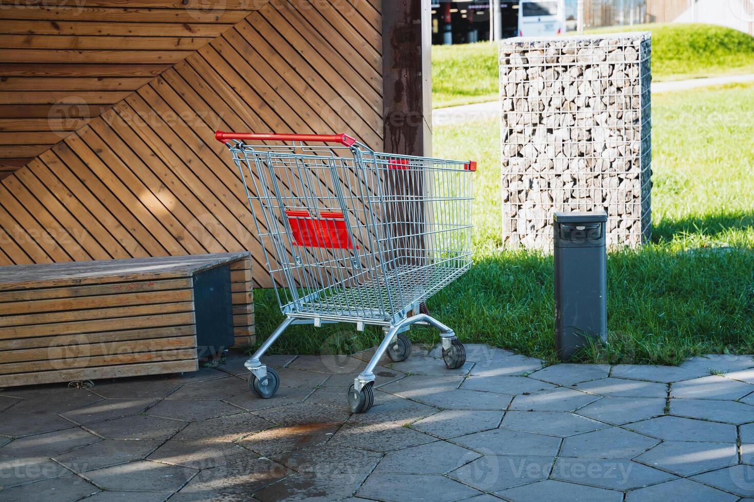 An empty shopping cart is currently parked on the sidewalk right in front of a wooden bench, creating a scene that captures the essence of everyday life in an urban environment photo