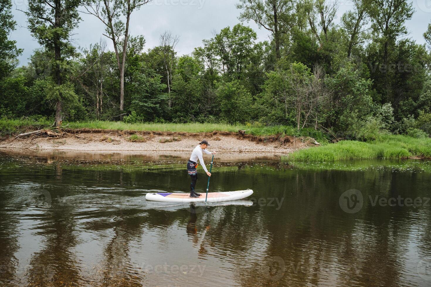 The man finds immense happiness paddle boarding in a tranquil river, surrounded by lush greenery under a blue sky, feeling a deep connection with the natural beauty surrounding him photo
