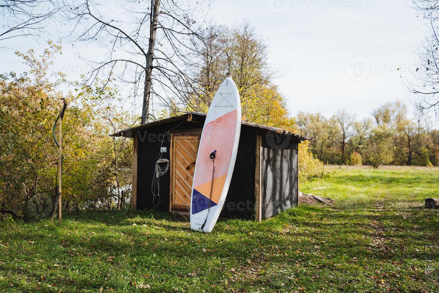 A colorful surfboard leans against a rustic shed, adding to the tranquil scene surrounded by lush green grass, enhancing the peaceful atmosphere of this picturesque setting photo