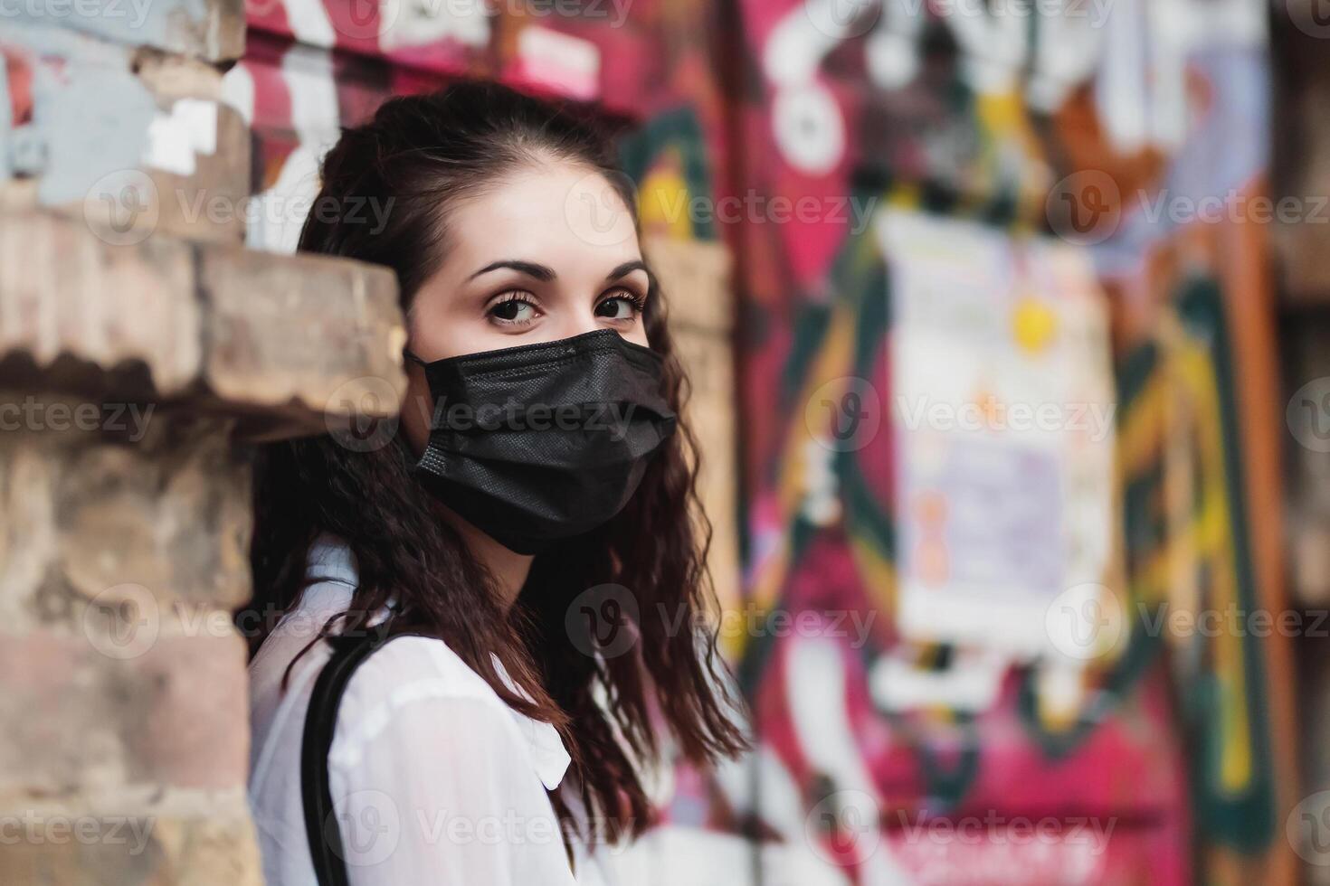 Young woman wearing black mask stands against urban graffiti wall photo
