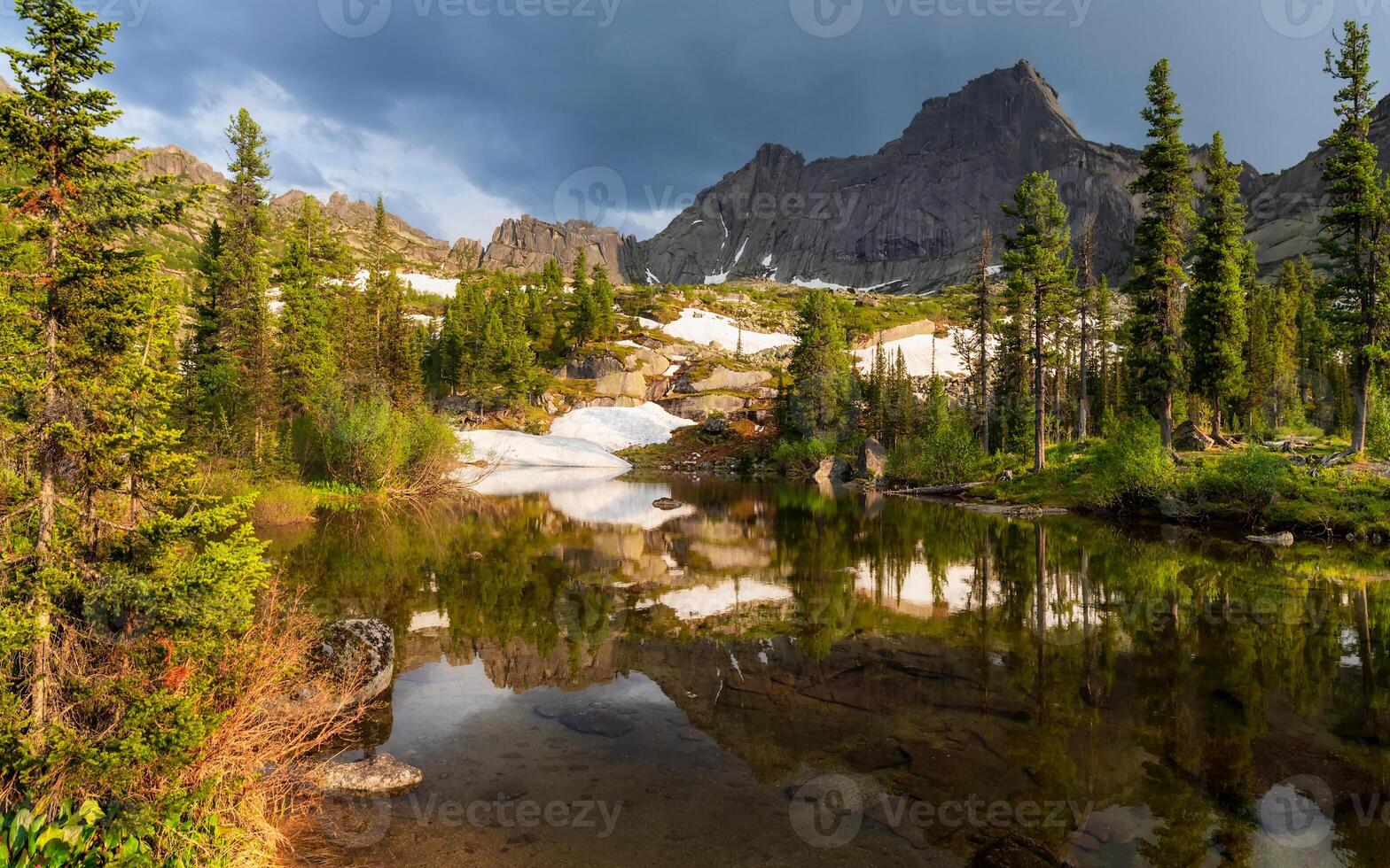 Taiga summer landscape. The purest mountain lake in the forest. Reflection of trees in the water. Sunny afternoon on the shore of a mountain lake, the remnants of snow on the slopes are melting. photo