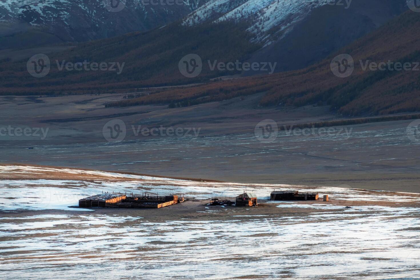 Small house, a kennel and a cattle pen at the foot of a snow-covered mountain in Altai on the bank of a frozen river. photo