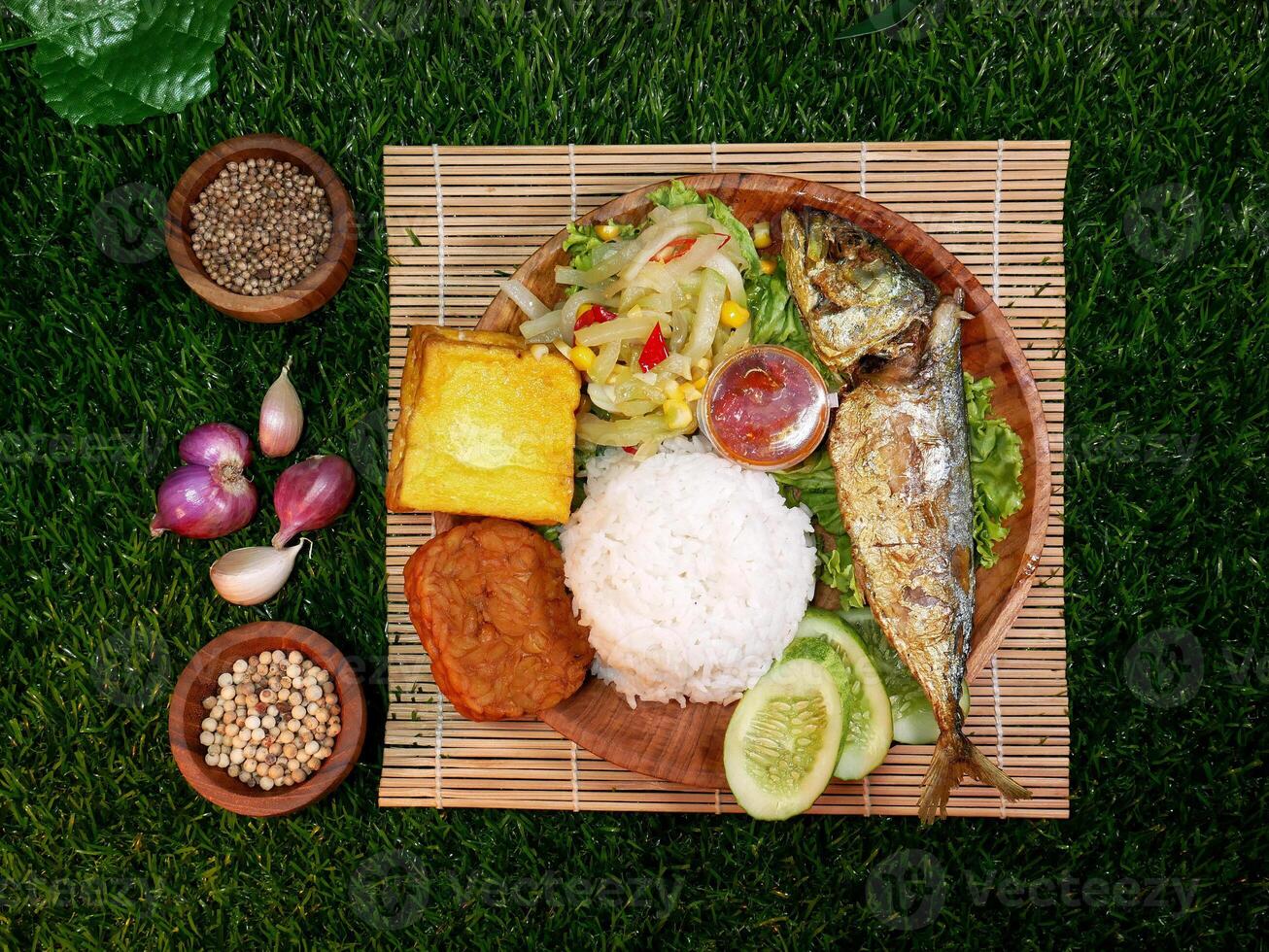 Top view, complete Mixed Rice or Nasi Campur served with white rice, fried fish, tempeh, tofu, fresh vegetables, sambal, and spices on a bamboo mat. photo