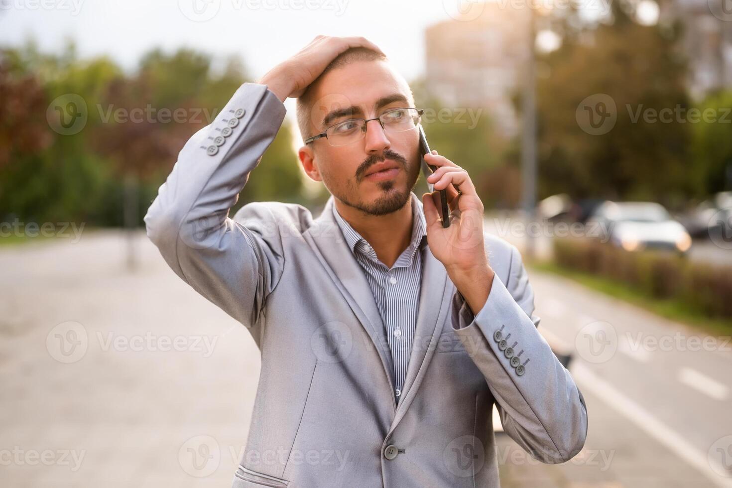 Distraught businessman is talking on phone. Young businessman standing in the street and using smartphone. photo