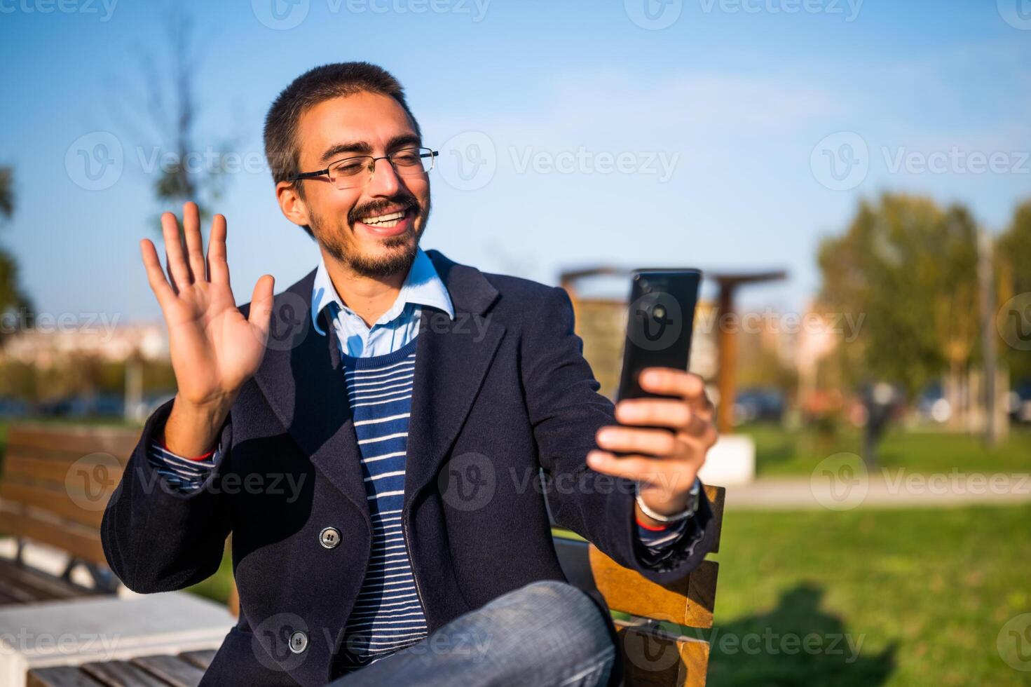 Happy businessman using mobile phone and waving while sitting on a bench in the park photo