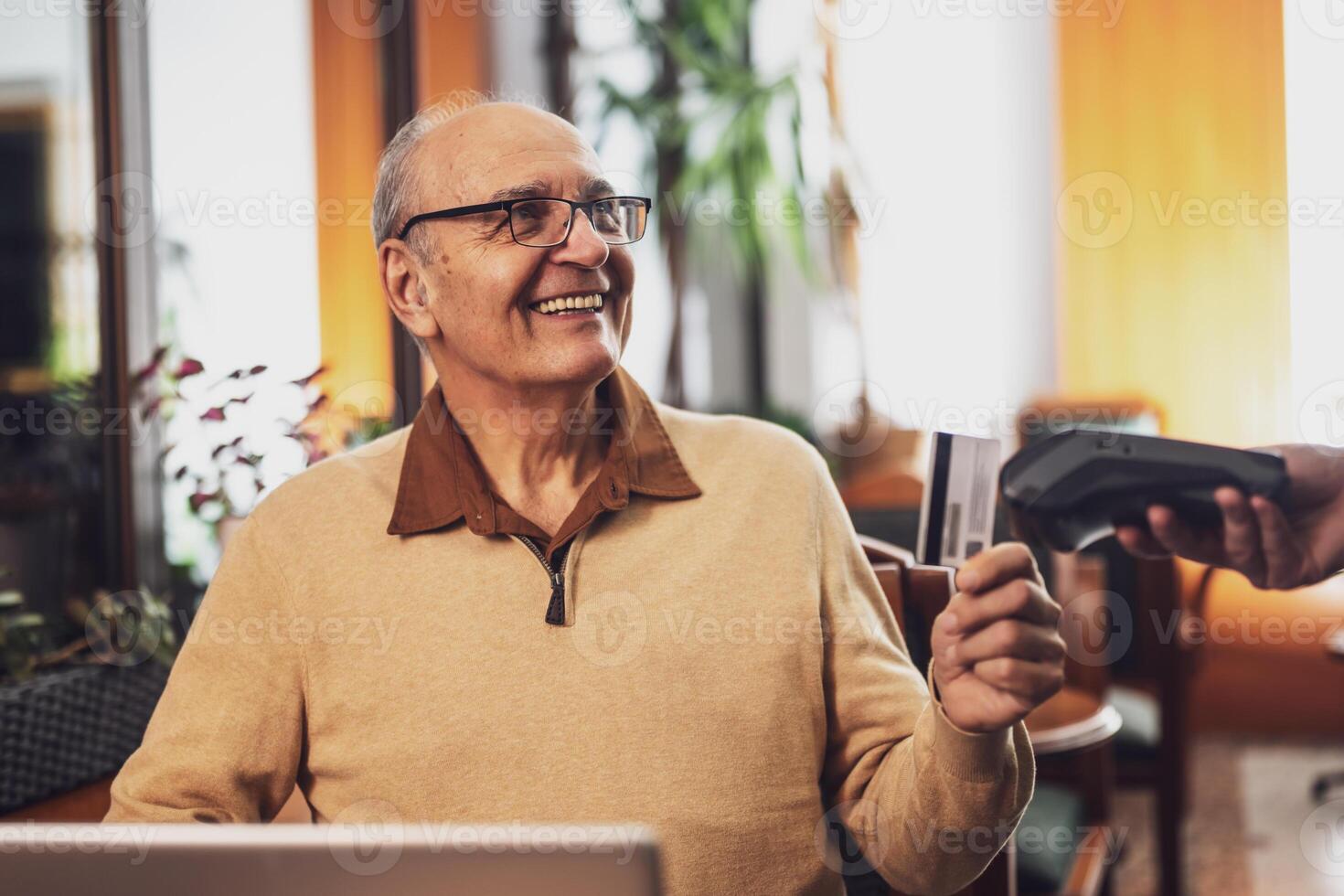 Cheerful senior man with eyeglasses in casual clothing making a contactless payment with credit card while sitting in the cozy city cafe at evening. photo