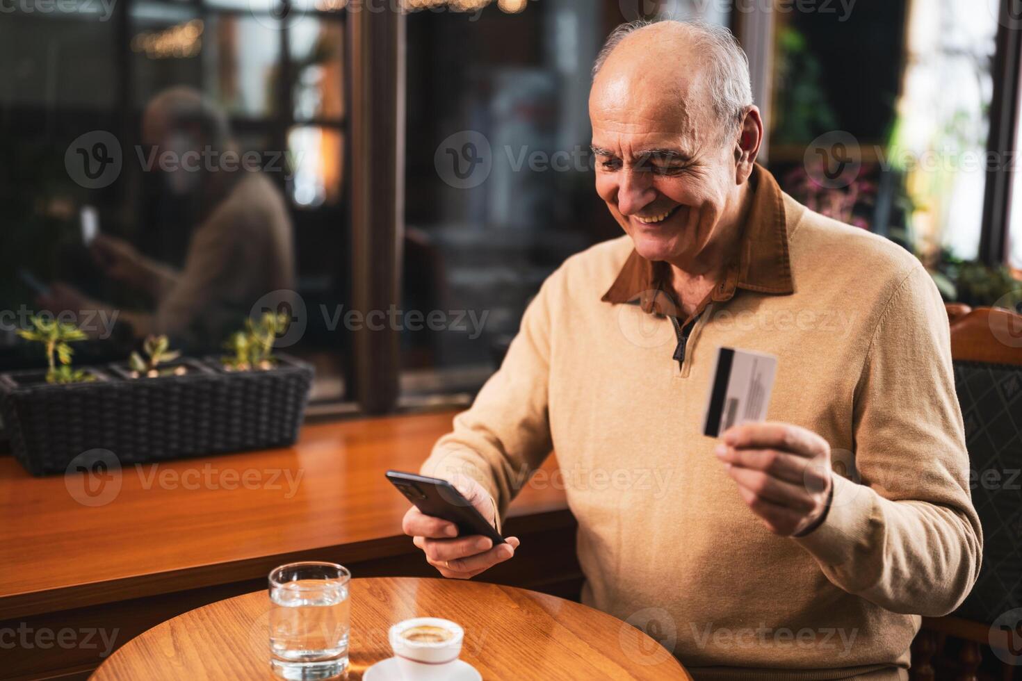 Happy senior man in casual clothing using smart phone and credit card while enjoying drinking coffee in the cozy city cafe photo