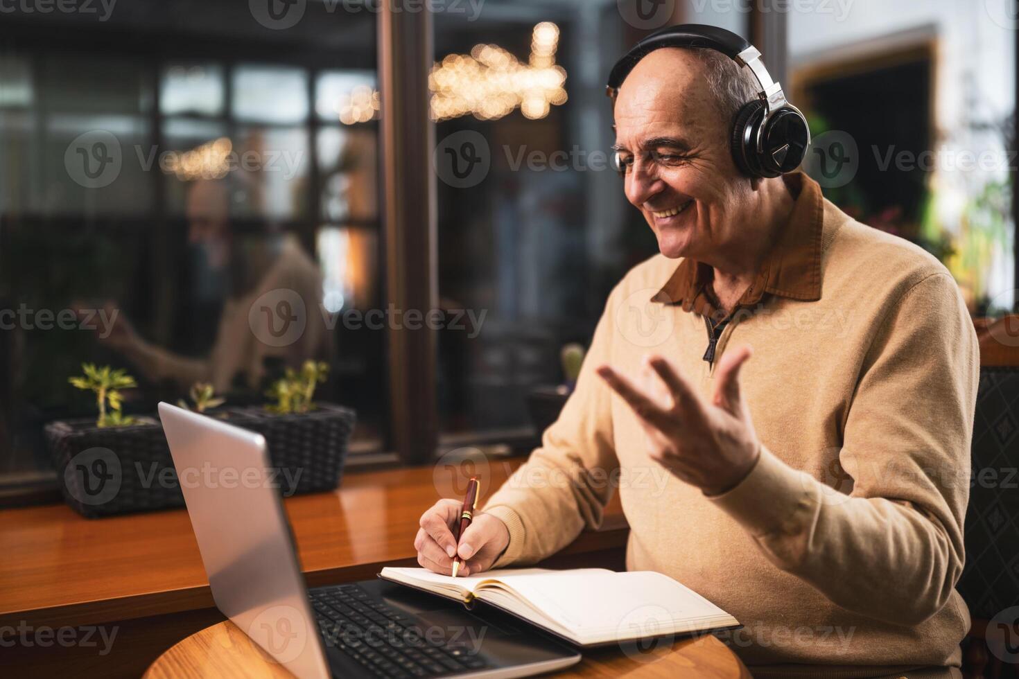 Cheerful senior man using wireless headphones, laptop and taking notes while attending online class. He is talking and gesturing during a call on computer in the cafe. photo