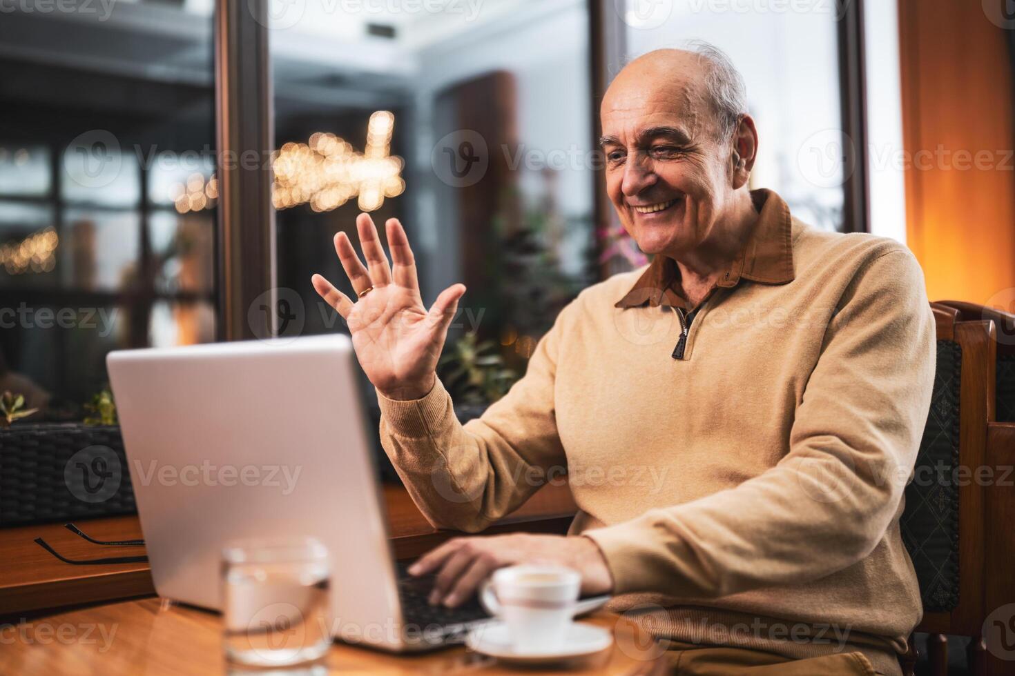 Cheerful senior man in casual clothing using laptop and waving during call while sitting in the city cafe photo