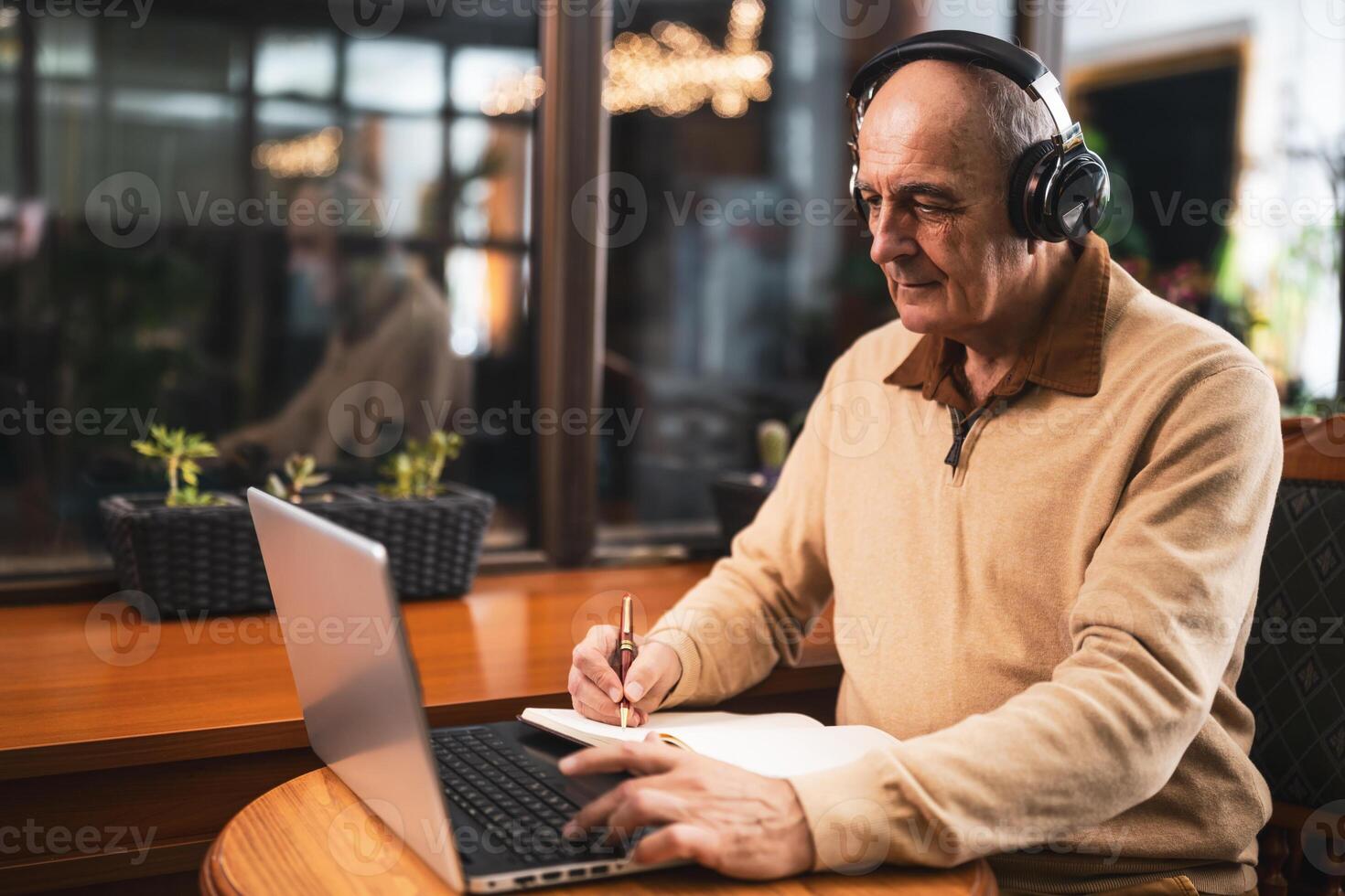 Focused senior man with wireless headphones learning online, writing notes and using laptop while sitting in the cafe. photo
