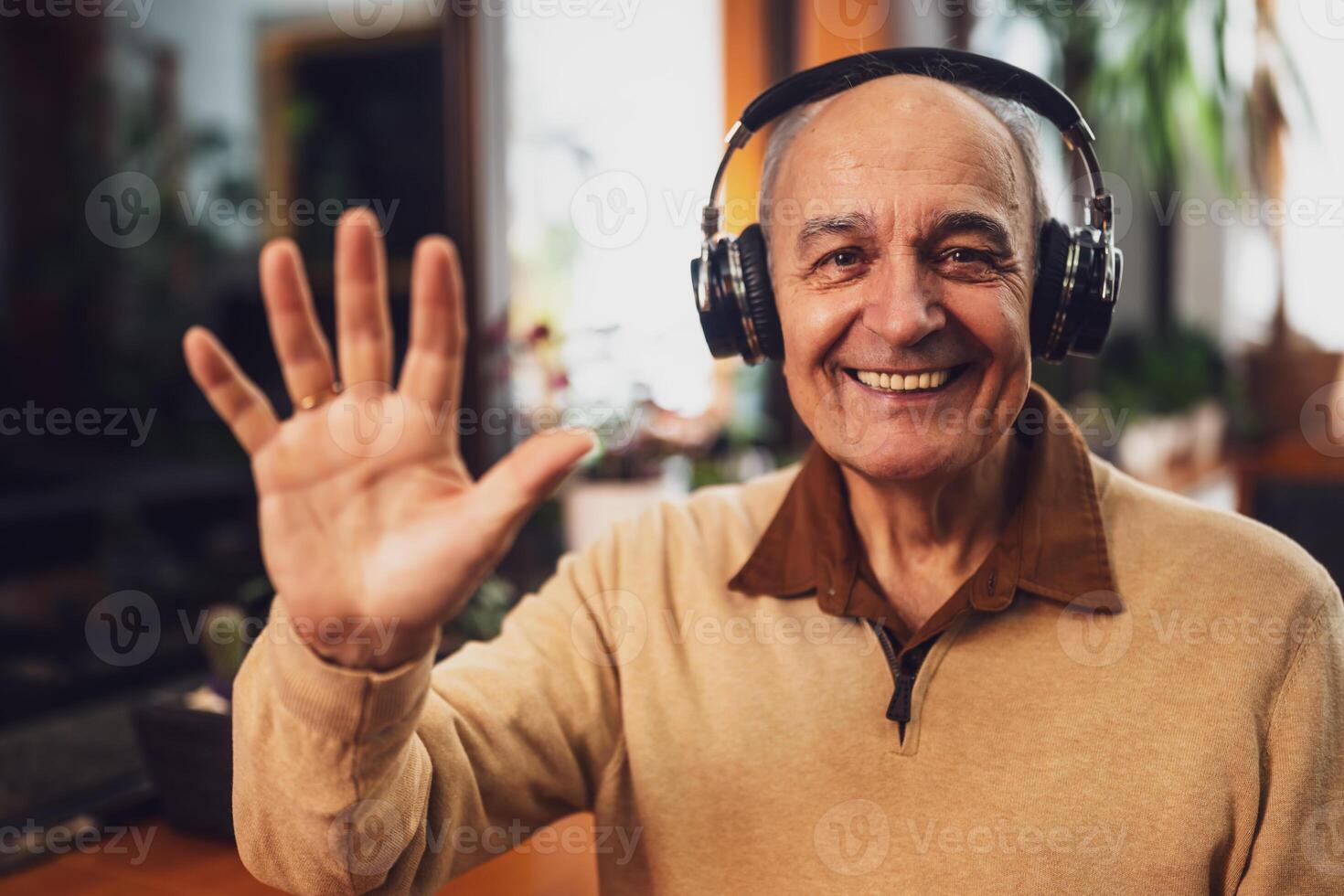 Portrait of smiling senior man in casual clothing with wireless headphones looking at camera and waving during a call on his laptop while sitting in the city cafe. photo