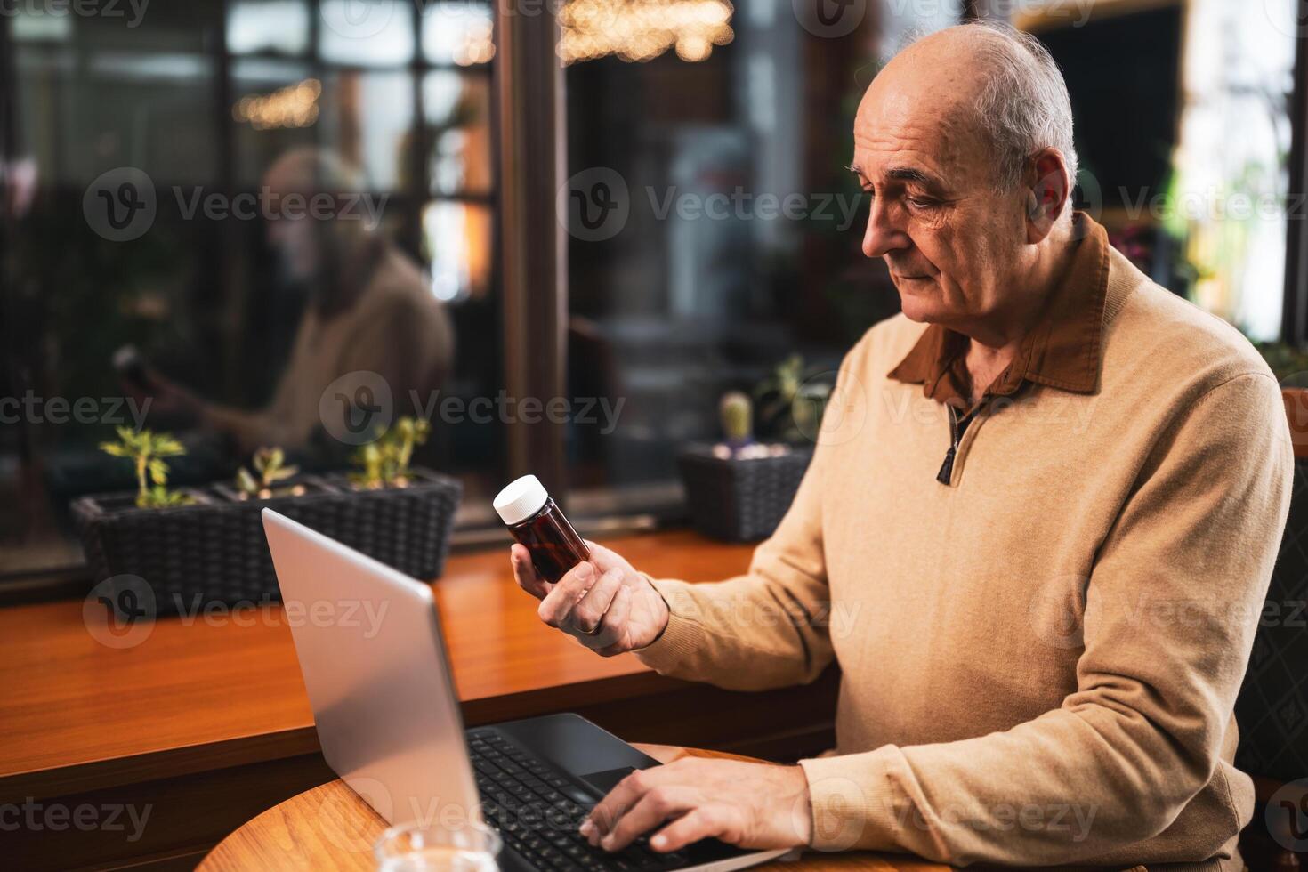 Senior man holding pill bottle and using laptop for searching more information about his prescription medicine while sitting in the cafe. photo