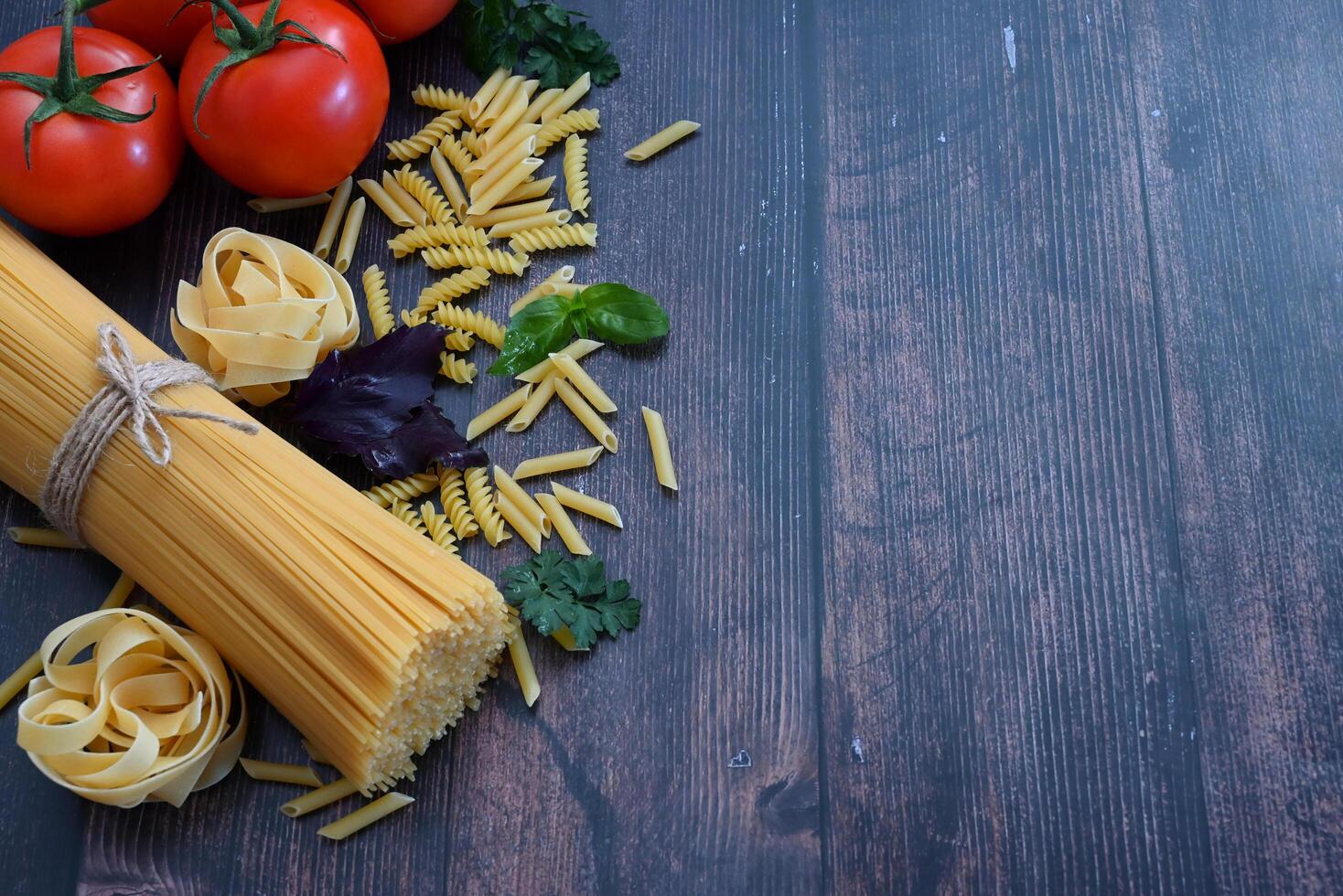 This photograph sets the stage for a pasta meal with ingredients like a bundle of spaghetti tied with a red ribbon, ripe tomatoes, chunks of cheese, garlic, fresh basil, and other additions photo