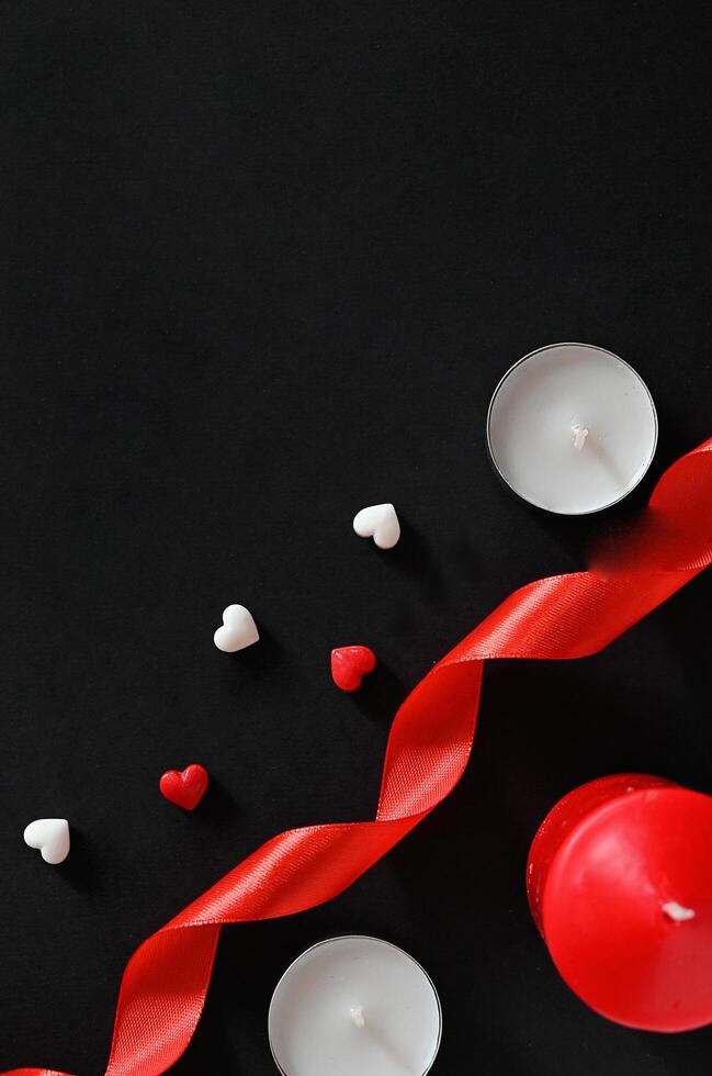 The picture shows a minimalist arrangement for a romantic occasion, using a white candle, a red candle, a red ribbon, and scattered heart-shaped candies in red and white, set against a plain photo