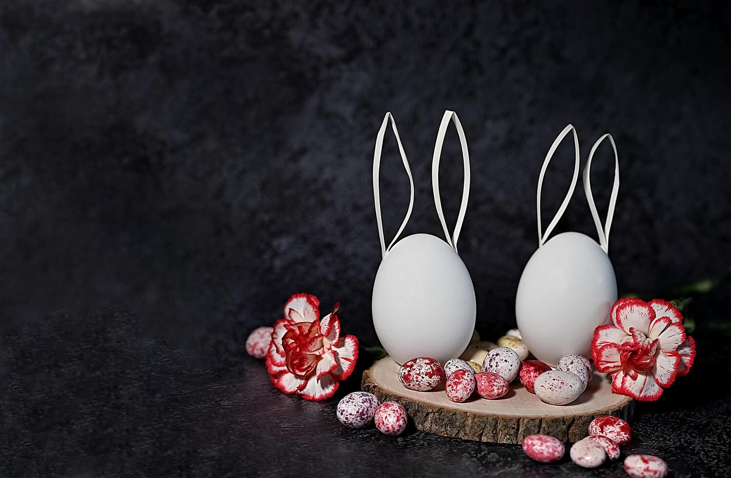 The photograph showcases a minimalist Easter decoration featuring two white eggs with white wire bunny ears, placed on a wooden base with speckled stones and yellow flowers. photo