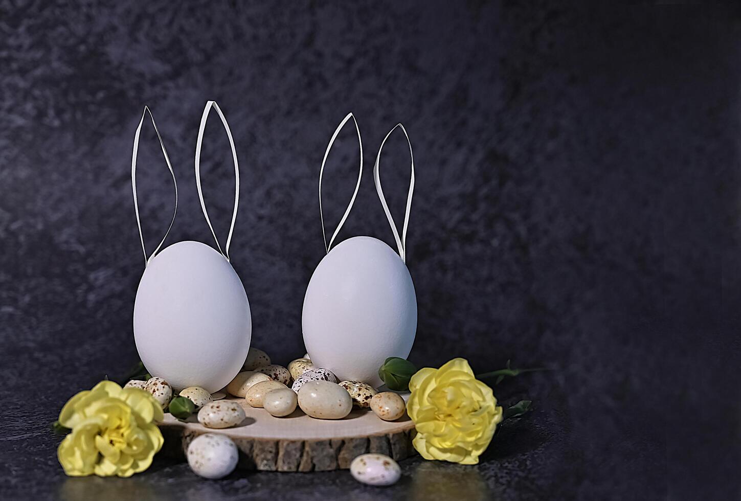 This image features two white eggs with paper bunny ears, arranged on a rustic wooden slice alongside small speckled stones and yellow flowers. photo